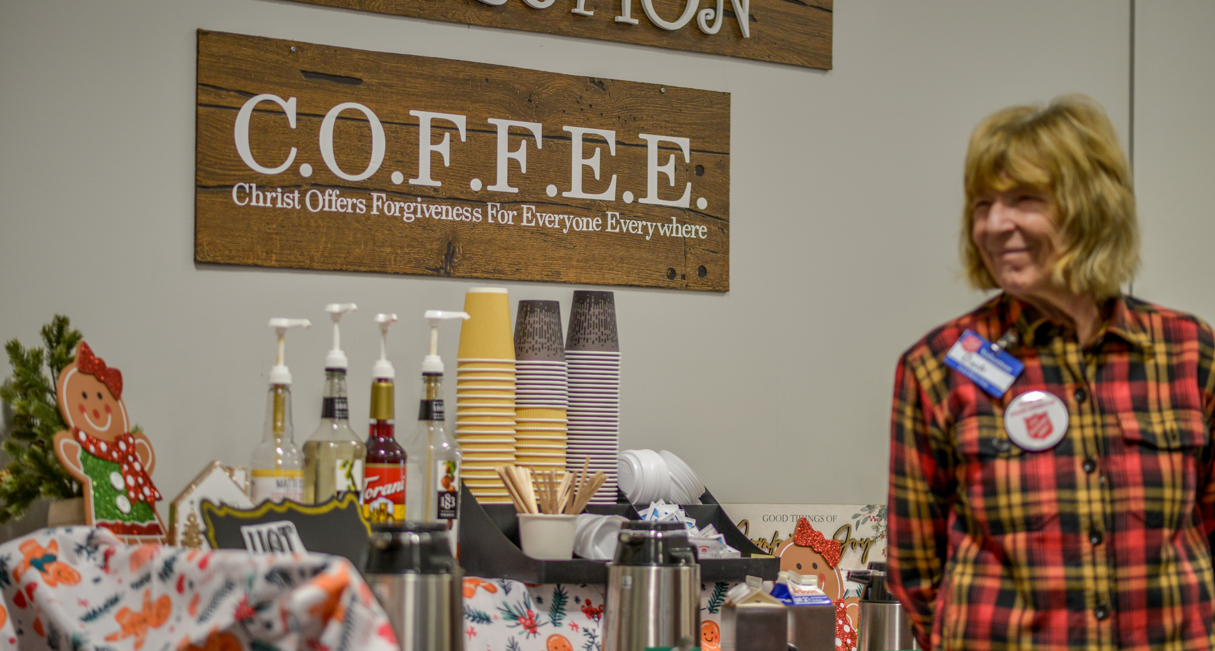 A smiling volunteer in a red plaid shirt stands beside a coffee station with syrup bottles, paper cups, and a wooden COFFEE sign with festive holiday decorations.