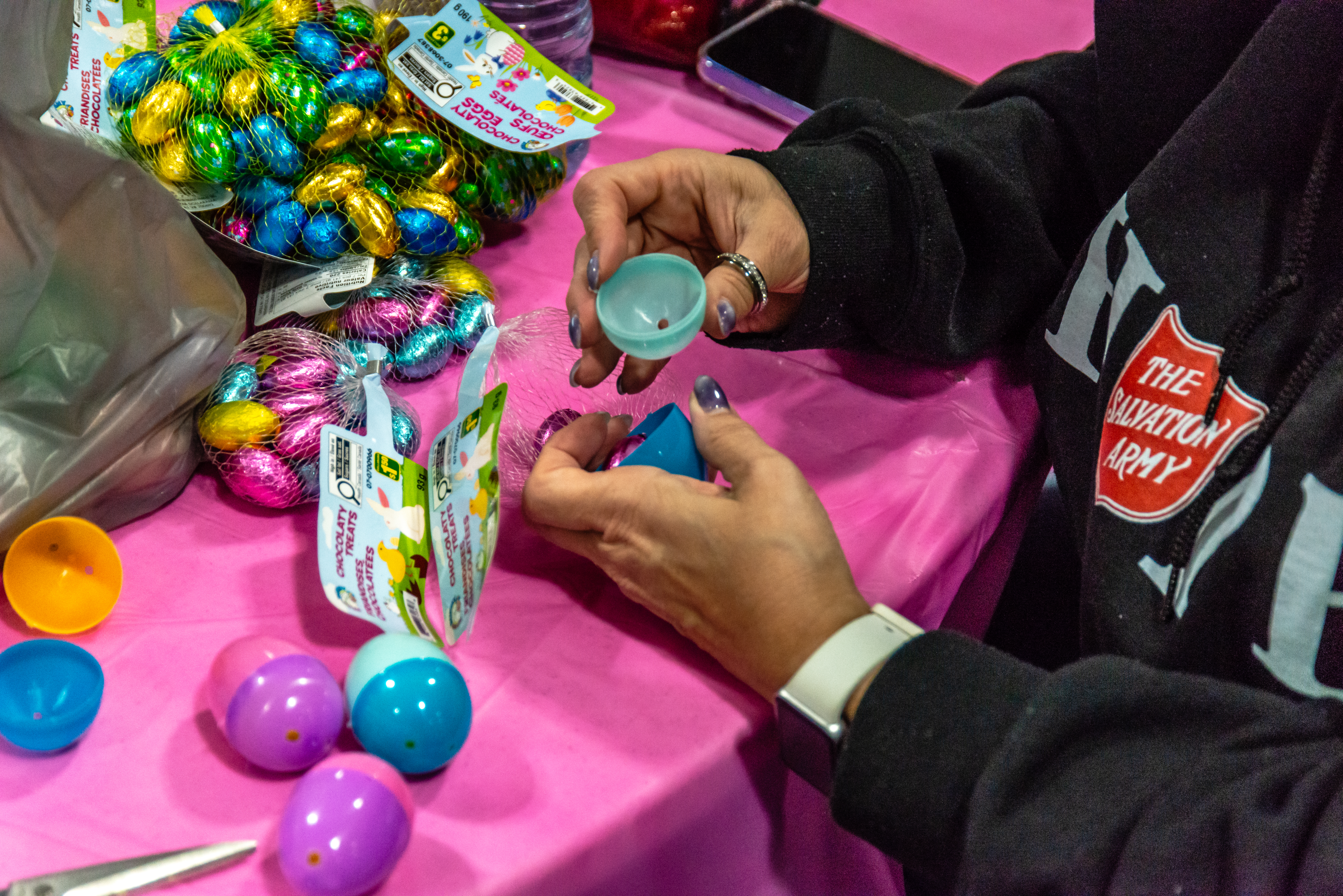 Hands filling colorful plastic Easter eggs with treats at a Salvation Army family event, with foil-wrapped chocolates scattered on a bright pink tablecloth.