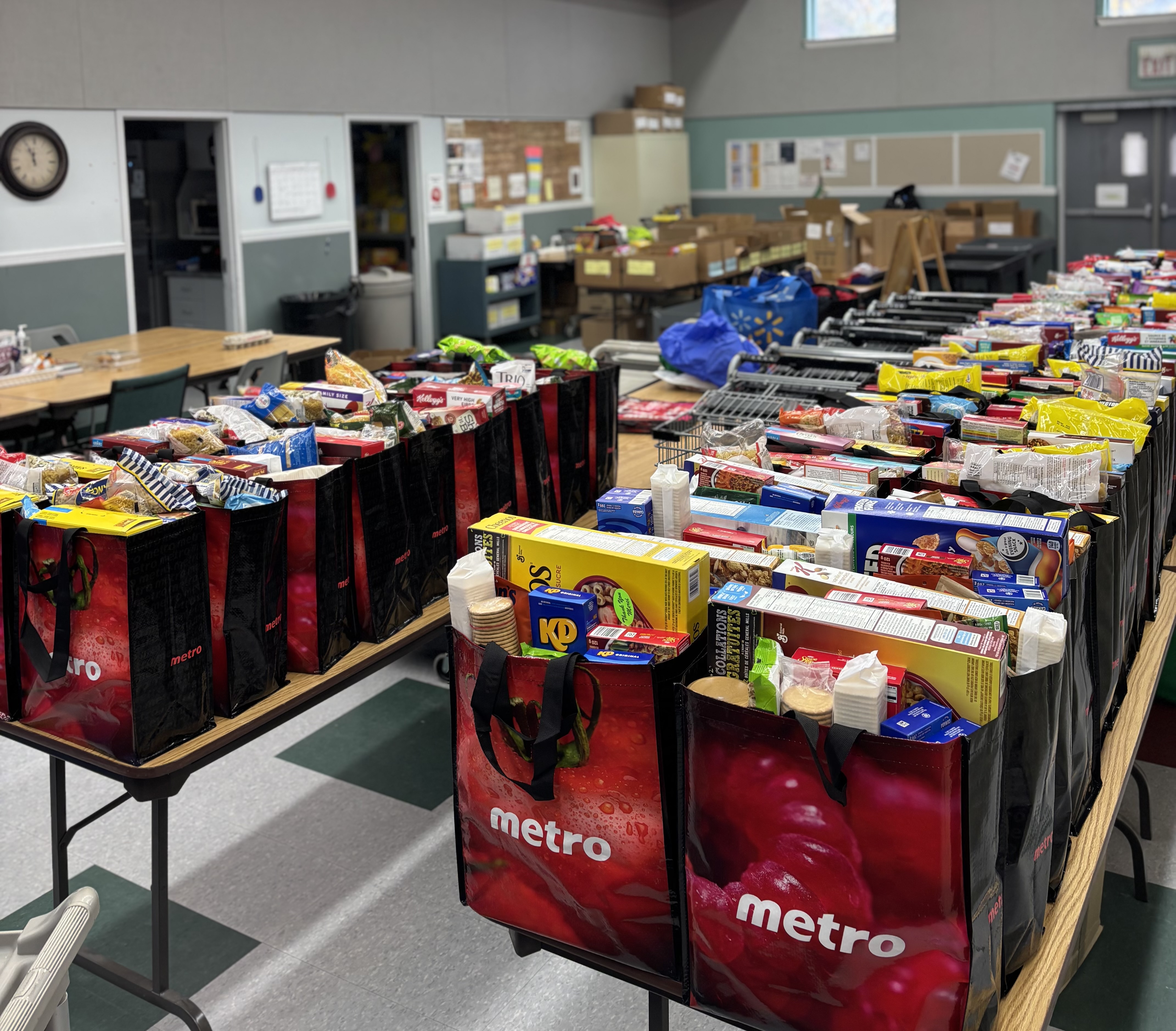 Rows of reusable bags placed on tables inside a room, each filled with groceries such as boxed pasta, cereal, canned goods, and snacks, prepared and organized for food bank distribution.