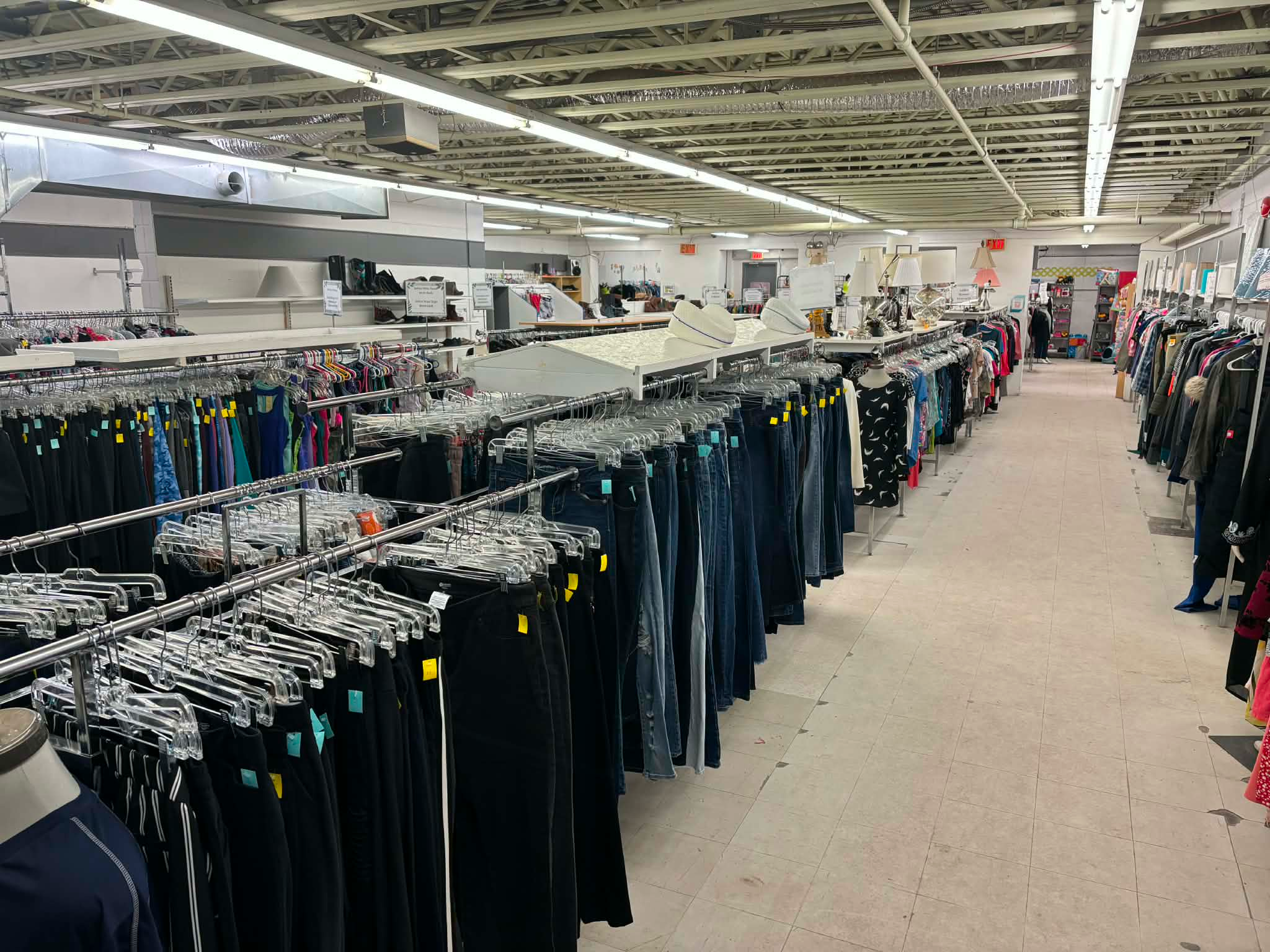 Interior of a thrift store with long rows of clothing racks filled with assorted garments, organized by type and color, with shelves and displays of additional items visible throughout the brightly lit space.