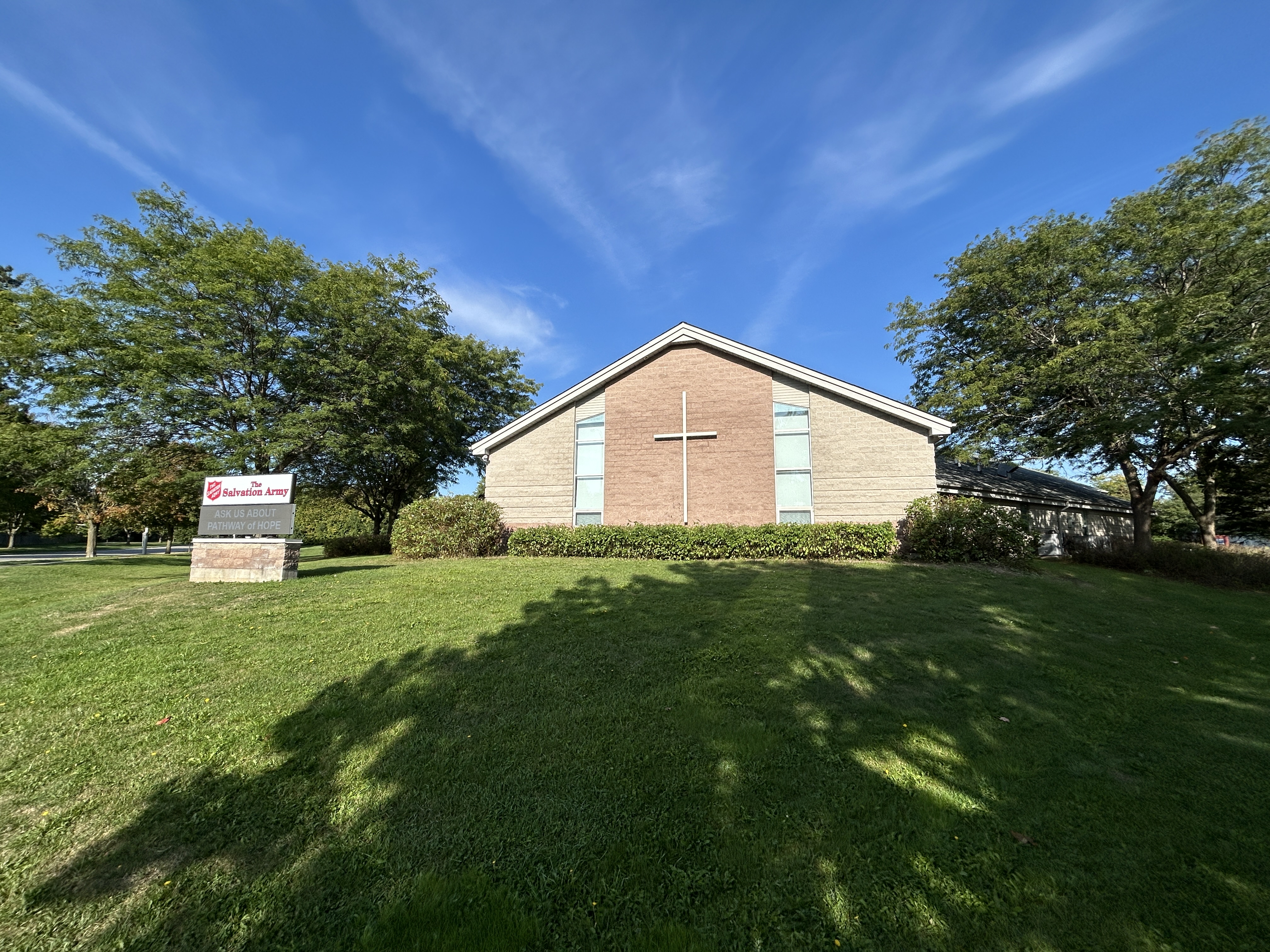 Exterior view of The Salvation Army Whitby Community Church building with a large cross on the front facade, surrounded by a green lawn, trees, and a sign near the entrance under a clear blue sky.
