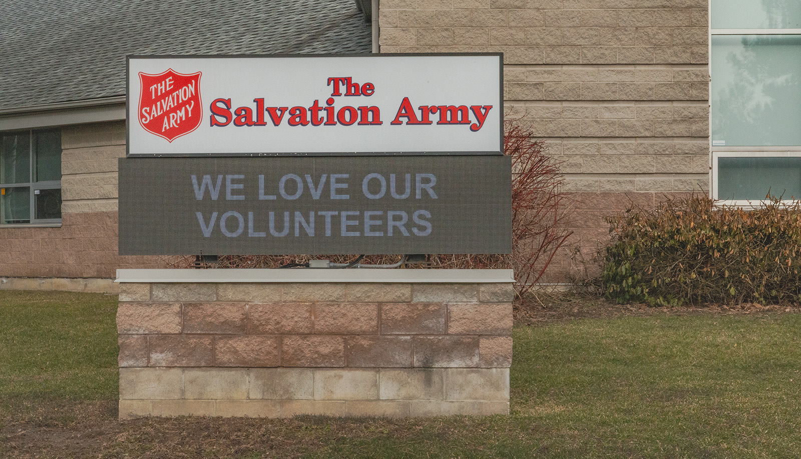 Outdoor Salvation Army sign mounted on a stone base in front of a building, displaying the message ‘WE LOVE OUR VOLUNTEERS’ beneath the red Salvation Army shield logo, with grass and shrubs surrounding the sign.