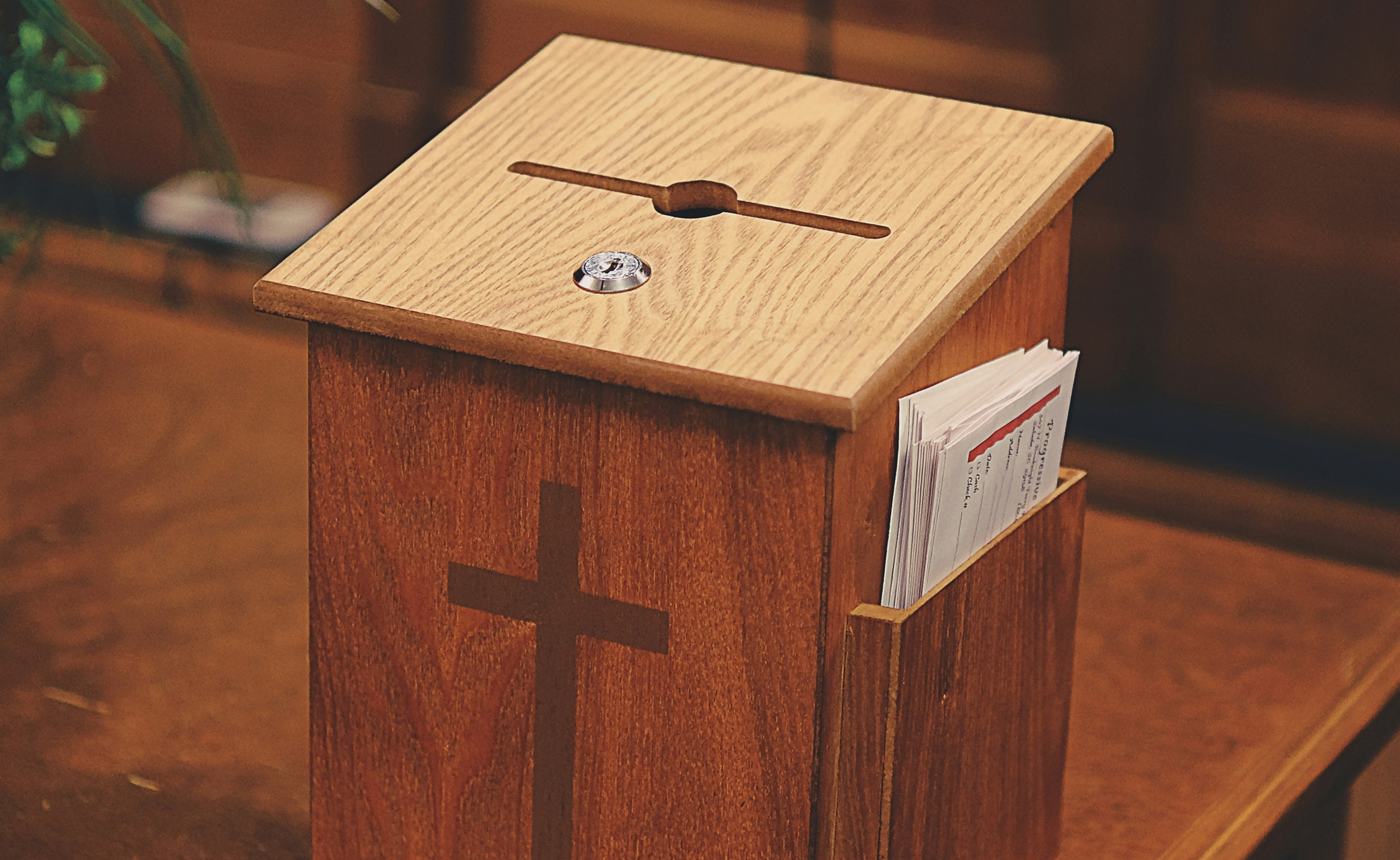 Wooden church offering box with a carved cross on the front and a slot on top, placed on a wooden surface with envelopes partially visible inside the side holder.