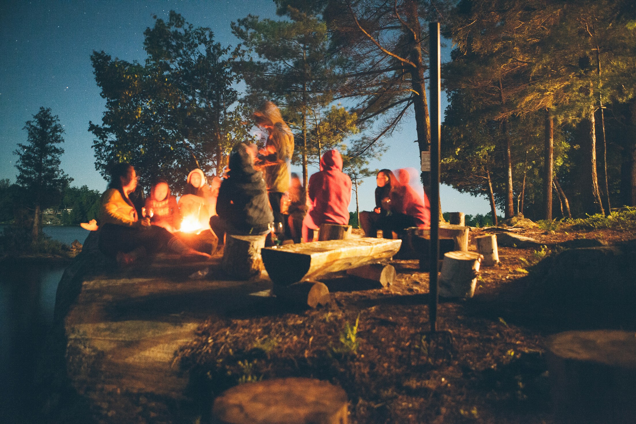 Group of children and leaders gathered around a campfire at night in a wooded outdoor setting, sitting on log benches with tall trees surrounding the area and a lake visible in the background.